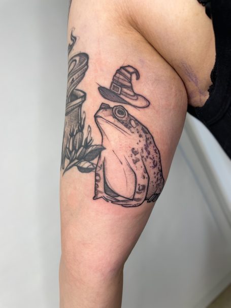 Ein Frosch mit Hut neben einer magischen Pflanze, tätowiert auf einem Oberarm.